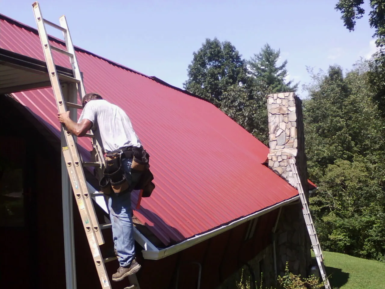 Roofer on a ladder beside a red metal roof and stone chimney — Soffit & Fascia Repair by Roofing Brothers in River Ranch